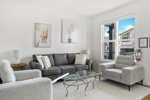 Un salon avec un canapé gris, une table basse en verre et deux fauteuils. Deux tableaux sont accrochés au mur, et une grande fenêtre offre une vue sur les bâtiments environnants.