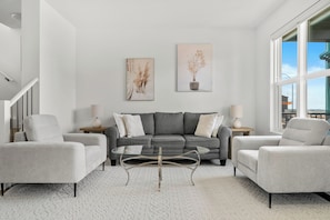 Un salon moderne avec un canapé gris, deux fauteuils, une table basse en verre et deux tableaux accrochés au mur.