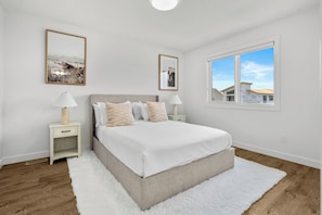 Une chambre à coucher avec un lit, deux tables de chevet, un tapis blanc, une fenêtre donnant sur un paysage agréable et deux tableaux encadrés accrochés au mur.
