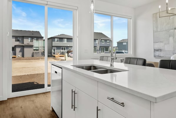 A modern kitchen with a large island, a double sink, and a view of a residential area through large windows.