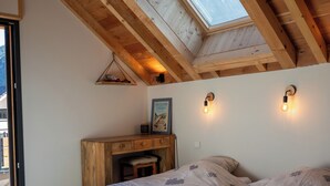 Une chambre avec un plafond en pente, des poutres en bois, un puits de lumière, deux lits avec une literie à motifs, un bureau en bois et un luminaire fixé au mur.