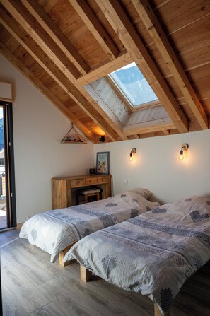 Ein Schlafzimmer mit schrägem Dach, Holzbalken, einem Oberlicht, zwei Betten mit gemusterten Bettdecken, einem Holztisch und einer wandmontierten Lampe.