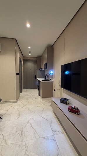 Camera d'albergo moderna con una grande TV a schermo piatto, un angolo cottura, un pavimento in marmo e un corridoio che conduce a un'altra stanza.
