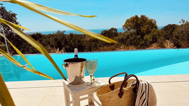 Une scène au bord de la piscine avec un seau en métal, deux verres, un sac en tissu tressé et une serviette à rayures posés sur un tabouret blanc.