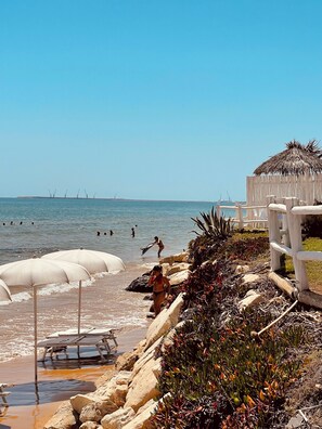 Une plage avec des parasols, une structure à toit de chaume et un terrain rocheux.