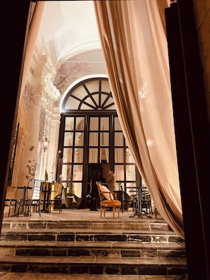 Uma escadaria imponente conduz a um espaço com uma ampla janela arqueada, detalhes decorativos elaborados e um candelabro.