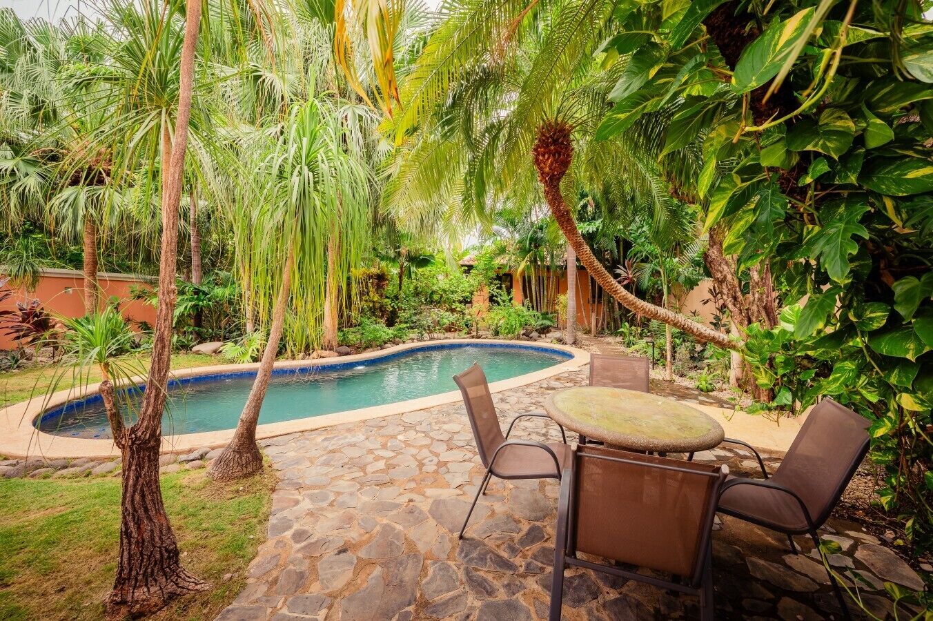 A pool surrounded by lush greenery and stone flooring, with a table and chairs nearby.