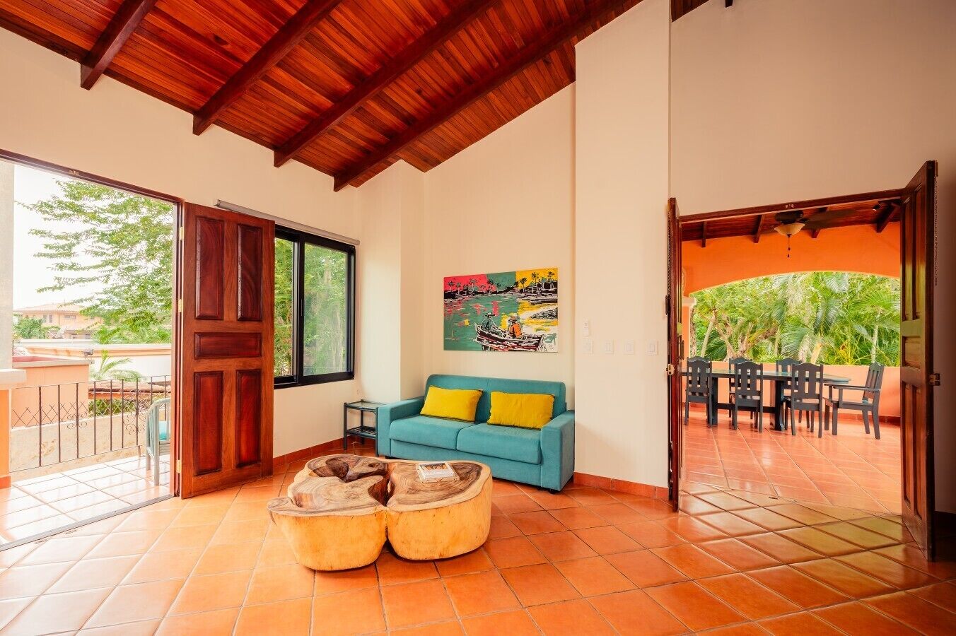 A living room with a wooden ceiling, a sofa with yellow cushions, a painting on the wall, and a dining area with a view of greenery.