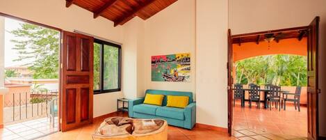 A living room with a wooden ceiling, a sofa with yellow cushions, a painting on the wall, and a dining area with a view of greenery.