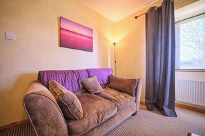 Uma sala de estar com sofá, luminária de chão, janela com cortinas e uma obra de arte na parede.
