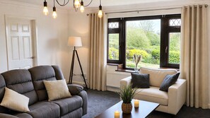 Ein Zimmer mit einem dunkelgrauen Sofa, einem weißen Sofa, einer Stehlampe, einem Tisch mit einer Kerze und Blick ins Grüne durch große Fenster.