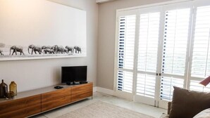 A modern living room with a flat-screen TV, a wooden cabinet, a large window with blinds, and a beige sofa. - Klein Champagne Guesthouse and Cottages (Franschhoek)