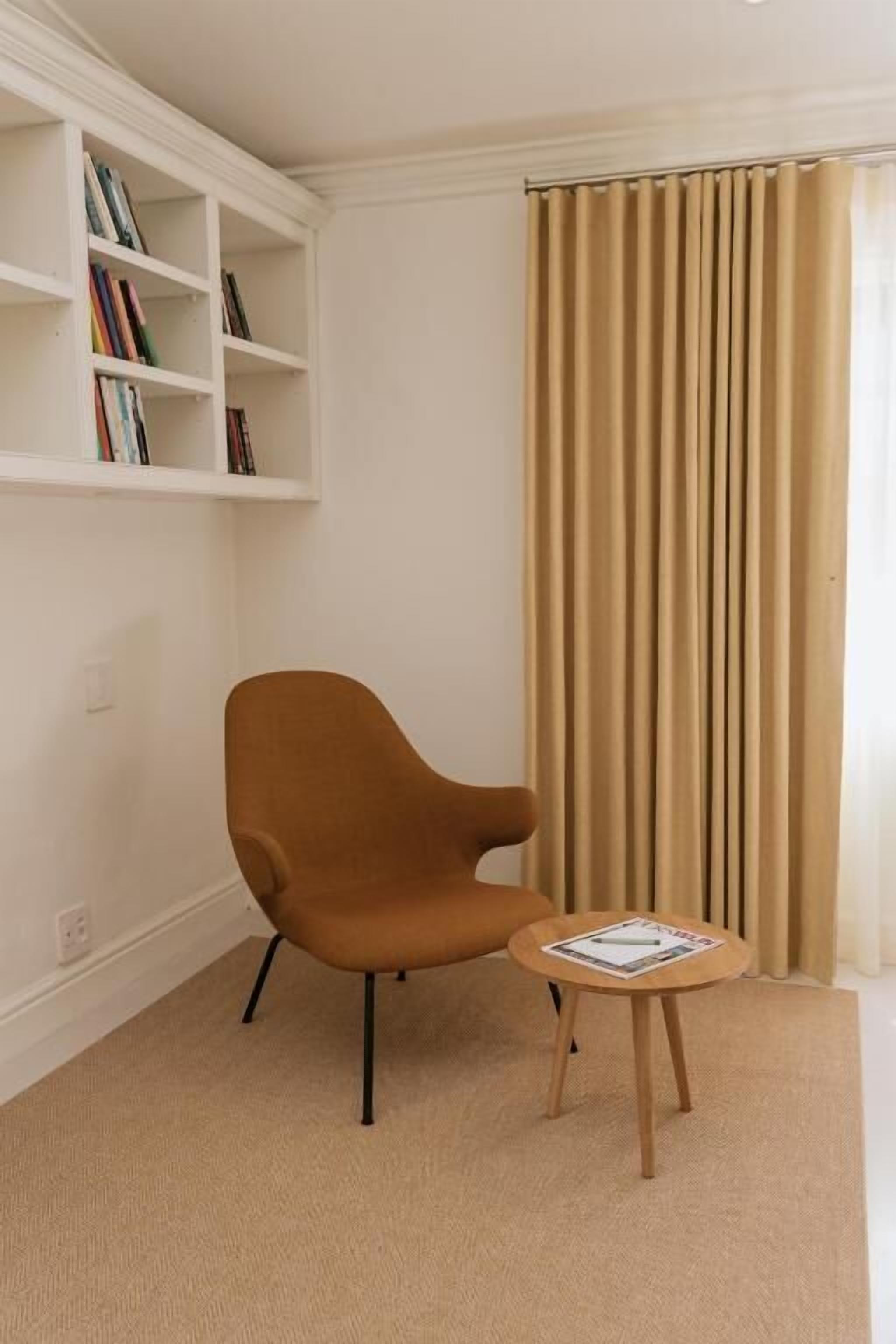 A room with a brown armchair, a small round table with a magazine, a bookshelf, and a window with beige curtains.