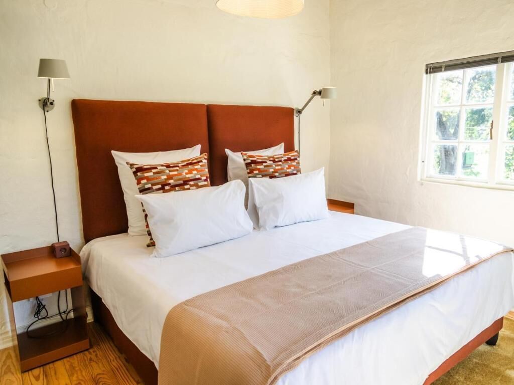 A bed with a brown headboard, white bedding, and patterned pillows. There are two bedside lamps, a small table with a radio, and a window with a view of greenery.