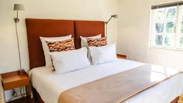 A bed with a brown headboard, white bedding, and patterned pillows. There are two bedside lamps, a small table with a radio, and a window with a view of greenery.