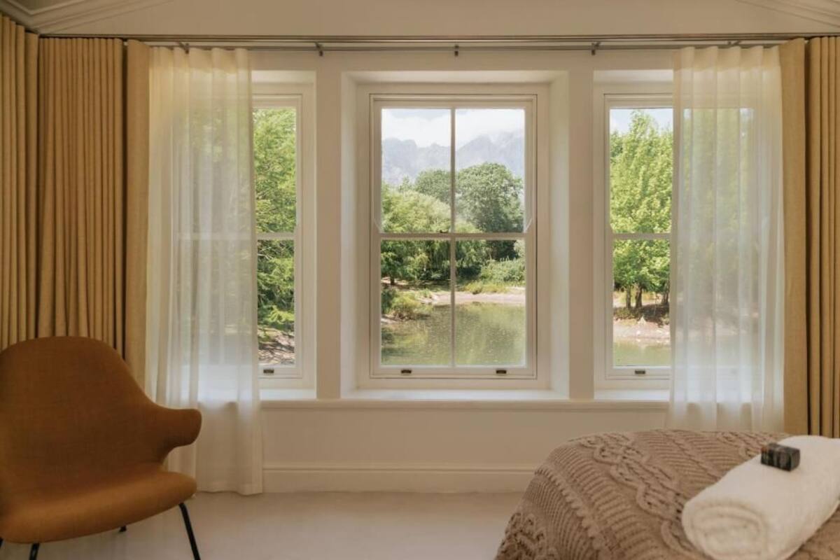 Una habitación de hotel con una cama, una silla y vistas a un lago y montañas a través de ventanas amplias.