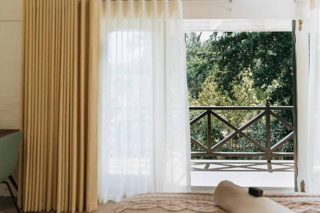 A room with a bed, a window with curtains, and a view of a balcony with a railing.
