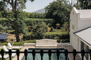 Una zona de piscina con una fuente de piedra, rodeada de exuberante vegetación y un edificio blanco.