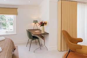 Habitación de hotel con cama, escritorio con silla, lámpara, florero con flores y ventana con vista a árboles.