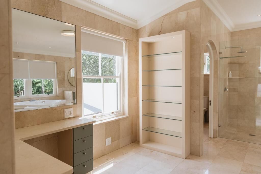 A modern bathroom with a large mirror, a window, a glass shelf unit, and a shower area.