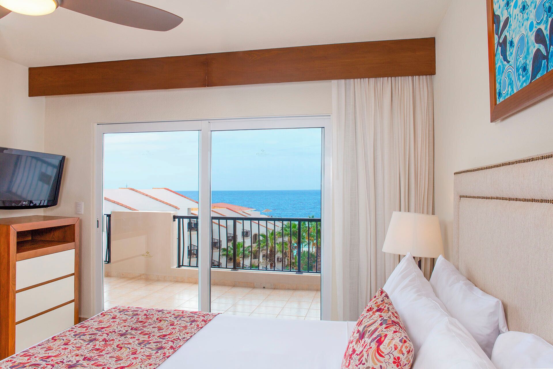 Habitación de hotel con cama, televisión, balcón con vista al mar y ventilador de techo.