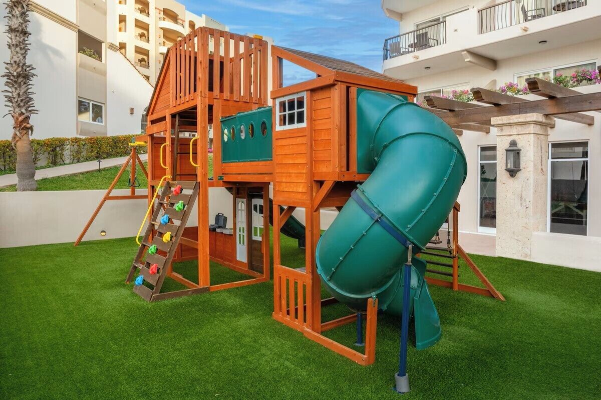 Une aire de jeux pour enfants avec un toboggan et une zone d’escalade, entourée de gazon synthétique et avec un bâtiment en arrière-plan.