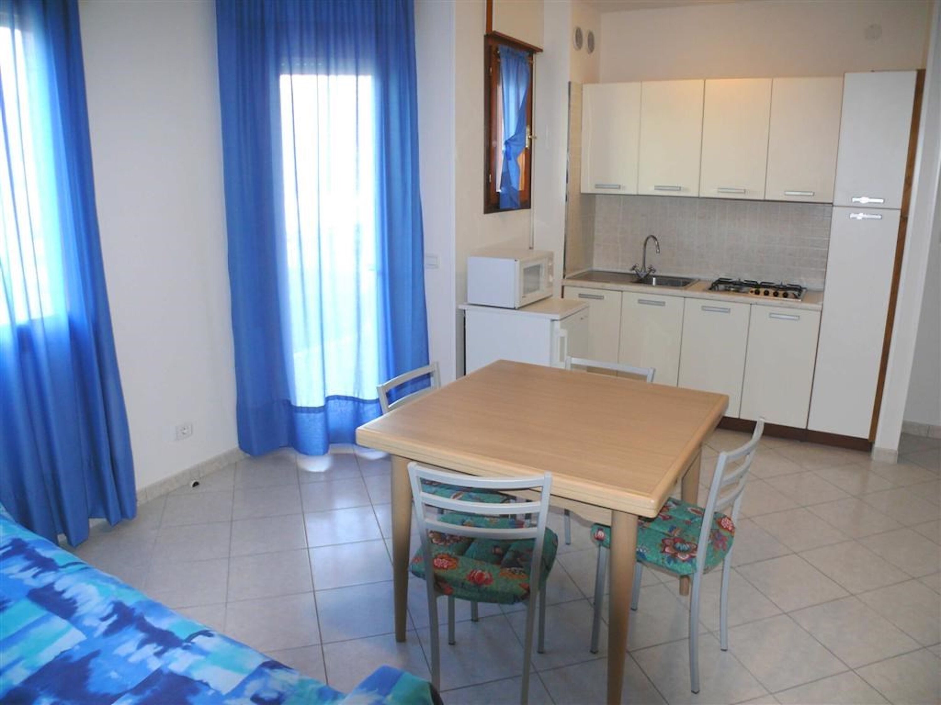 A small kitchen with white cabinets, a microwave, and a sink. There is a wooden table with two chairs, one with a floral cushion.