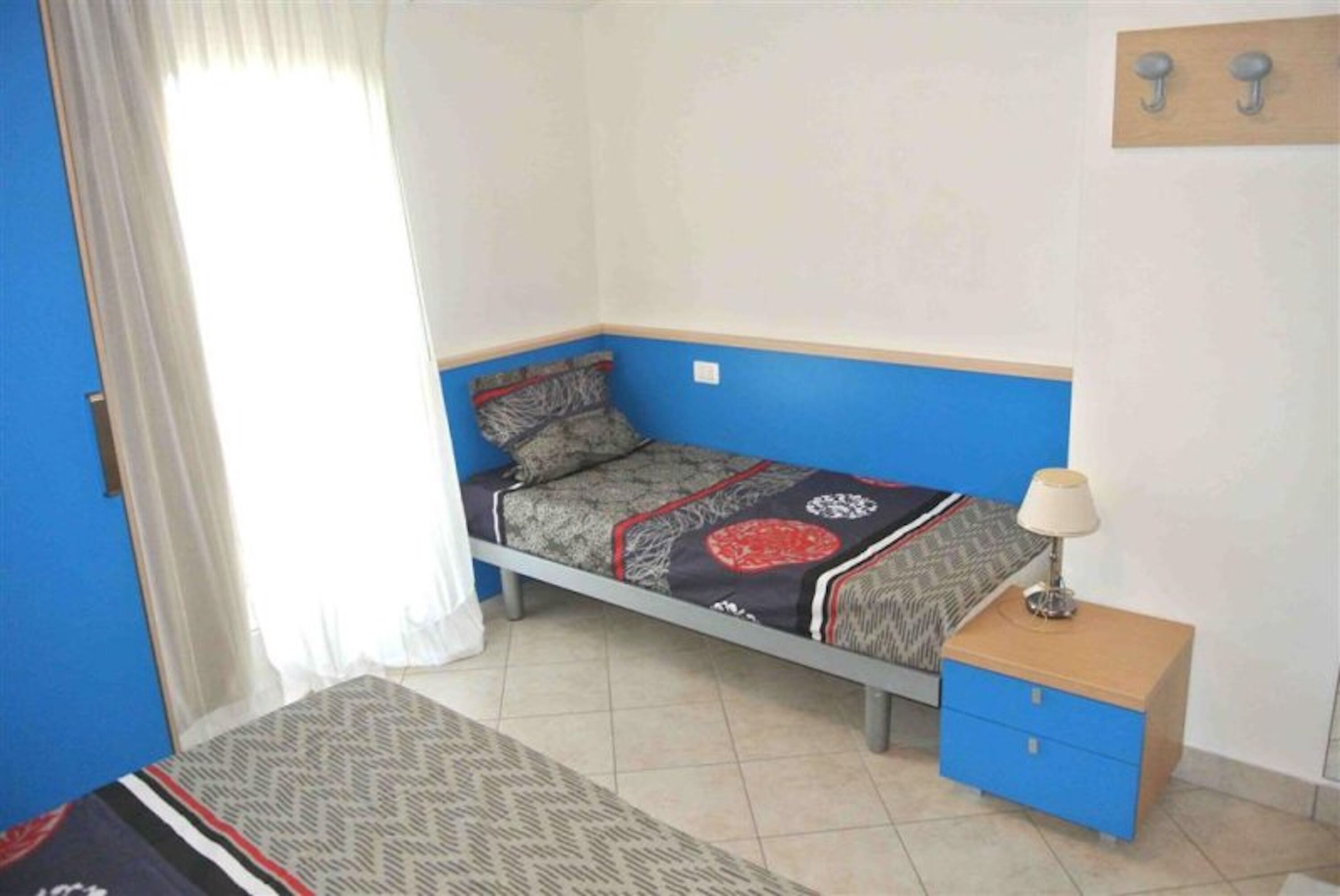 Un dormitori amb un llit, una nitzeta, una làmpada i una finestra amb cortines.
