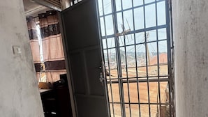 Ein Zimmer mit Fliesenboden, einer leicht geöffneten Tür und Blick durch ein vergittertes Fenster.