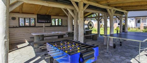 An outdoor recreational area with a foosball table, a ping pong table, and a television screen mounted on a wooden wall.