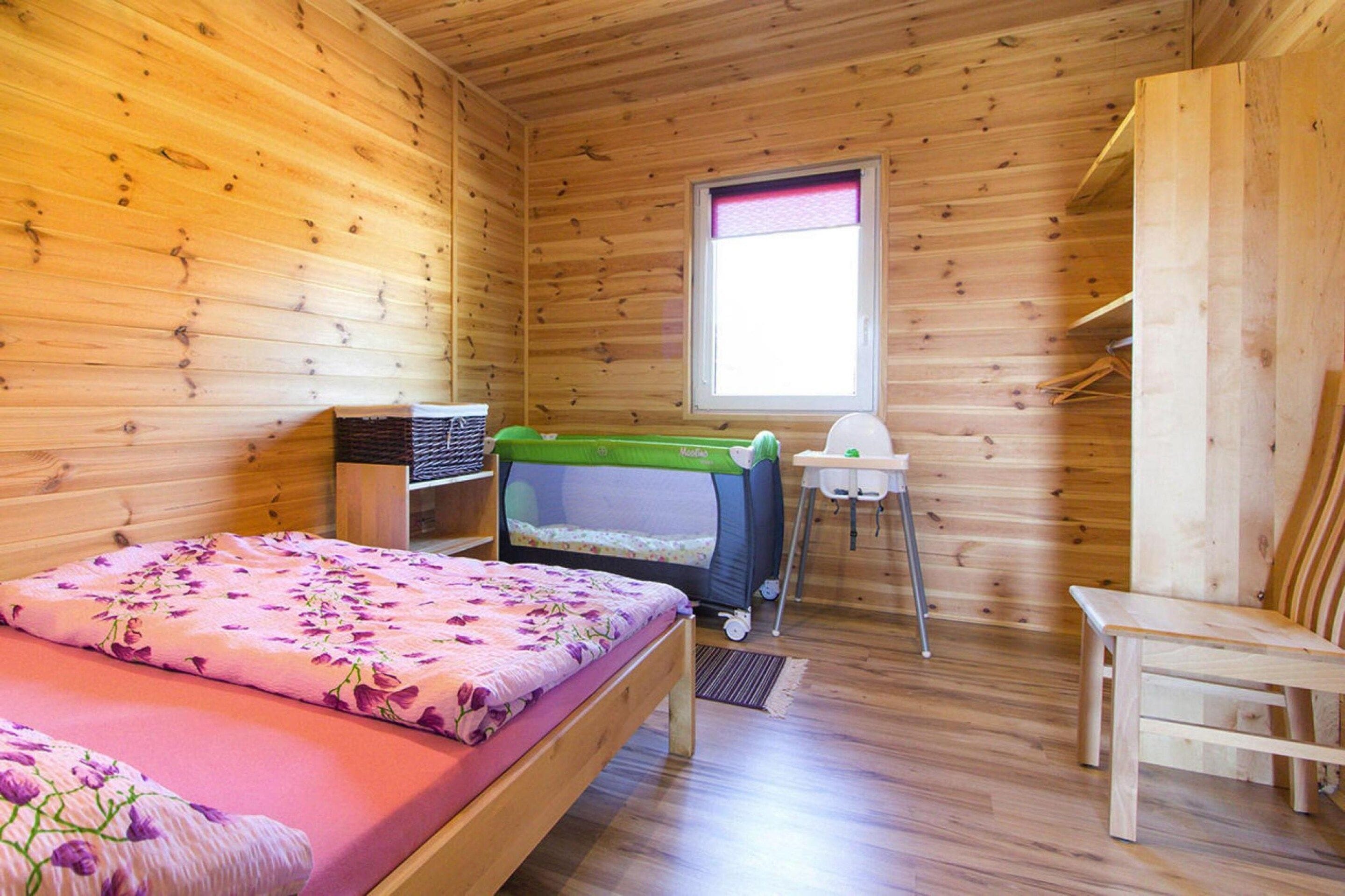 Ein Zimmer einer Holzhütte mit einem Bett, einem kleinen Tisch, einem grünen Kinderwagen und einem Fenster.
