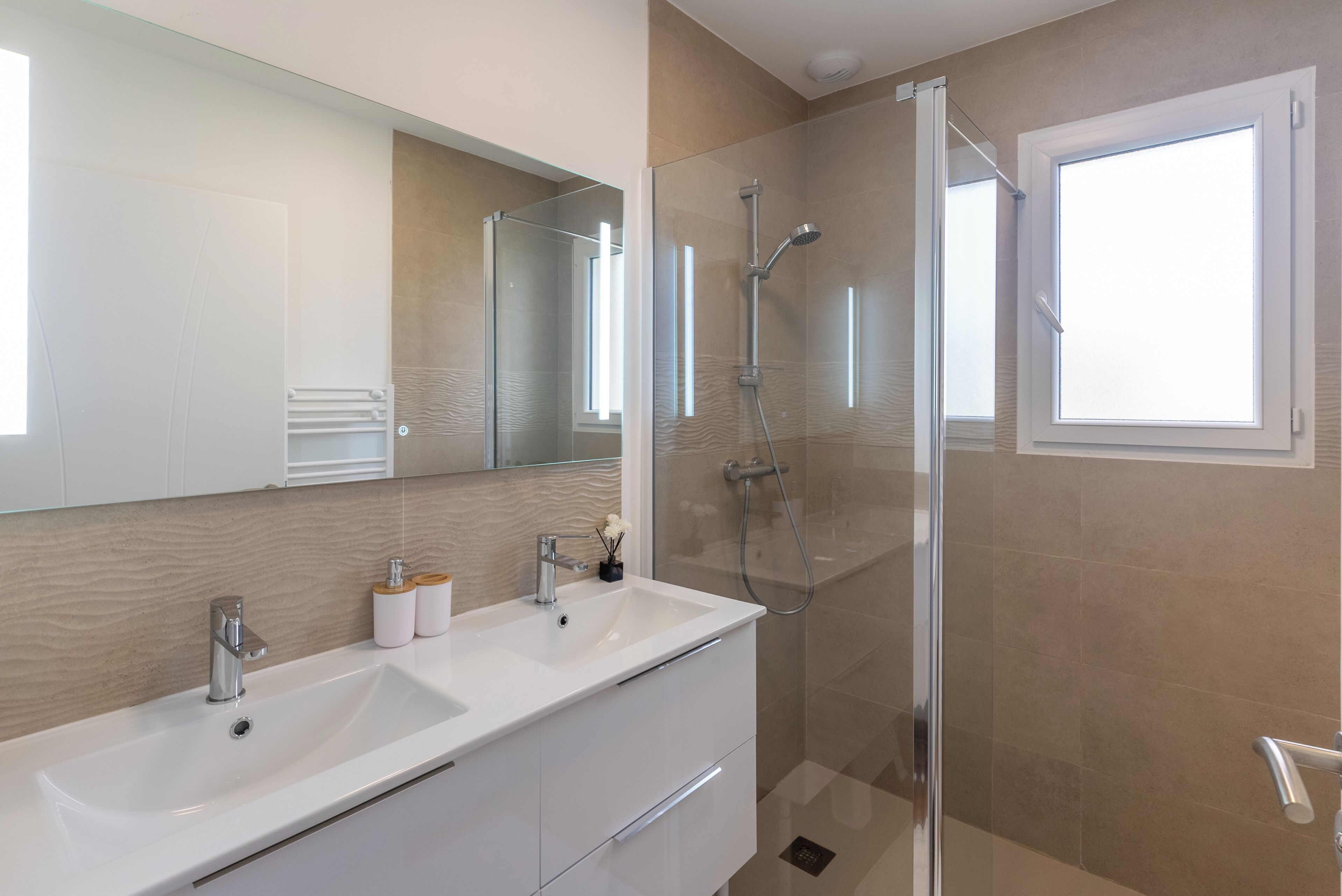A modern bathroom with a glass shower enclosure, a white sink with a mirror, and a window providing natural light.