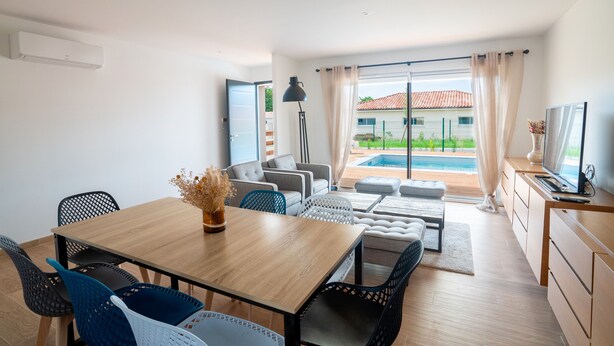 Una sala de estar moderna con una mesa de comedor de madera, sillas blancas y negras, un conjunto de sofá, un televisor y vista a una piscina.