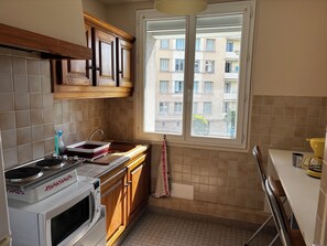 A kitchen with wooden cabinets, a microwave, a sink, and a window with a view of buildings. - Quiet apartment in Aubenas. 10 min walk from the Château. (Aubenas)