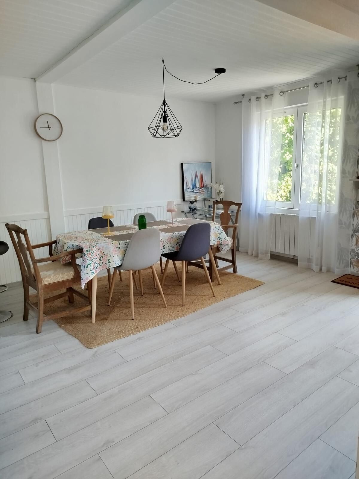 Een eetruimte met een tafel gedekt voor vier personen, een hanglamp, een klok aan de muur en een raam met gordijnen.