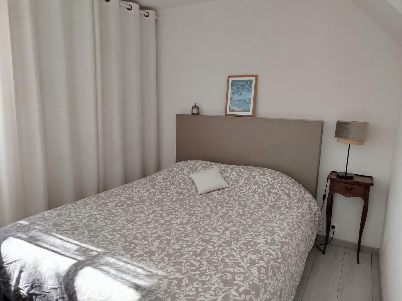 Un dormitorio con cama, mesita de noche, lámpara, un cuadro enmarcado en la pared y una ventana con cortinas.