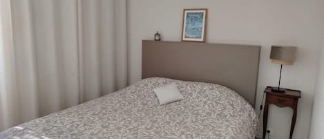 Un dormitorio con cama, mesita de noche, lámpara, un cuadro enmarcado en la pared y una ventana con cortinas.