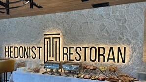 Montaje de buffet con pared de piedra de fondo y un letrero que dice 'HEDONIST RESTORAN'.