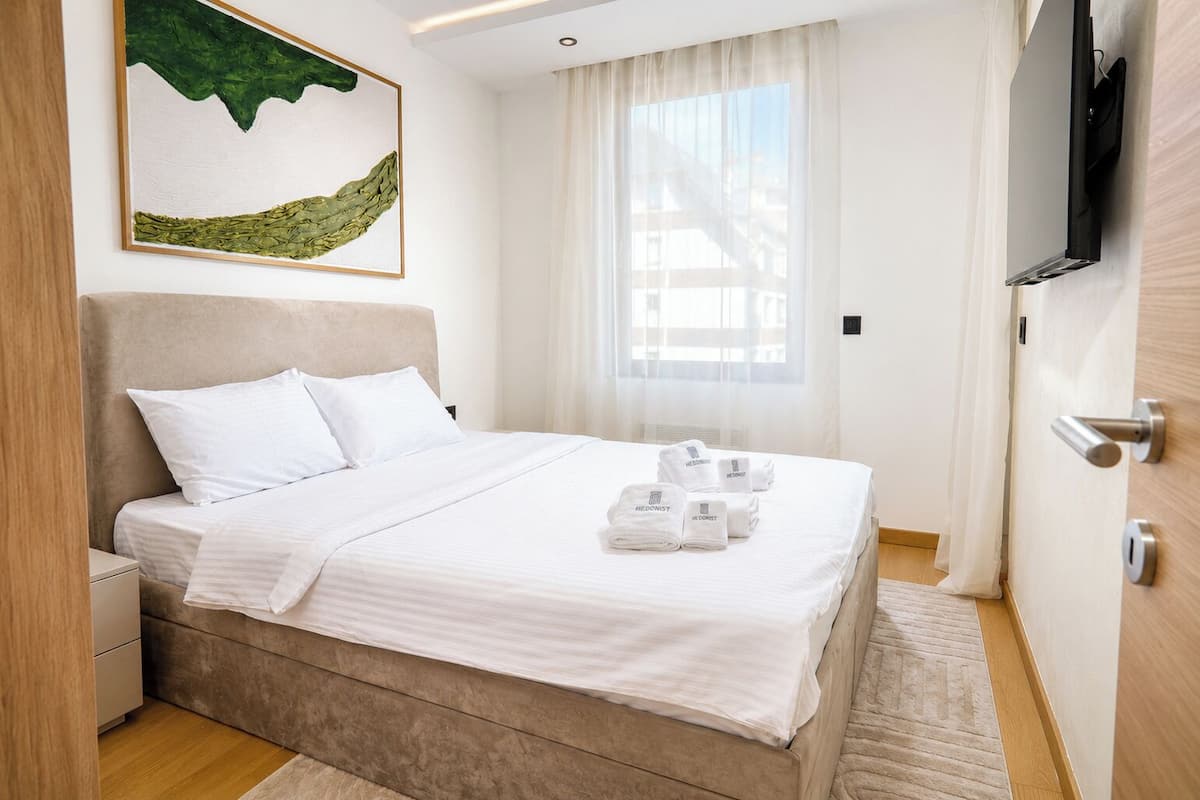 A neatly made bed with white linens and towels, a painting above it, and a television on the wall.
