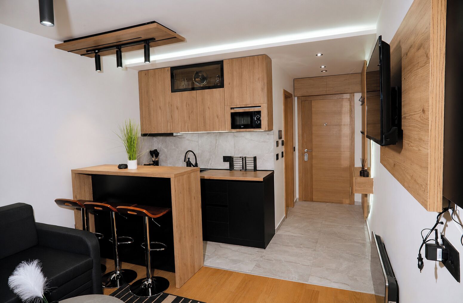 A compact kitchen with a microwave, a small sink, and a wooden countertop. There is a black chair with a white feather pillow, a small plant, and a black bar stool. A television is mounted on the wall.
