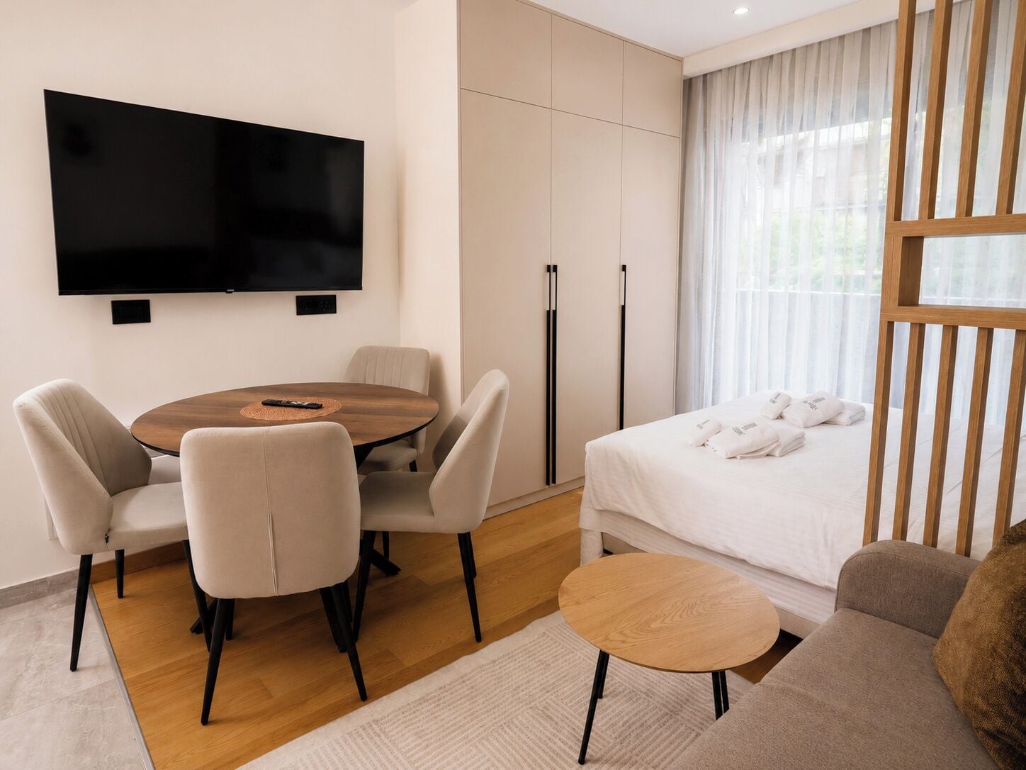 A modern hotel room with a flat-screen TV, a round dining table with chairs, a bed with white linens, a small round table, and a window with sheer curtains.