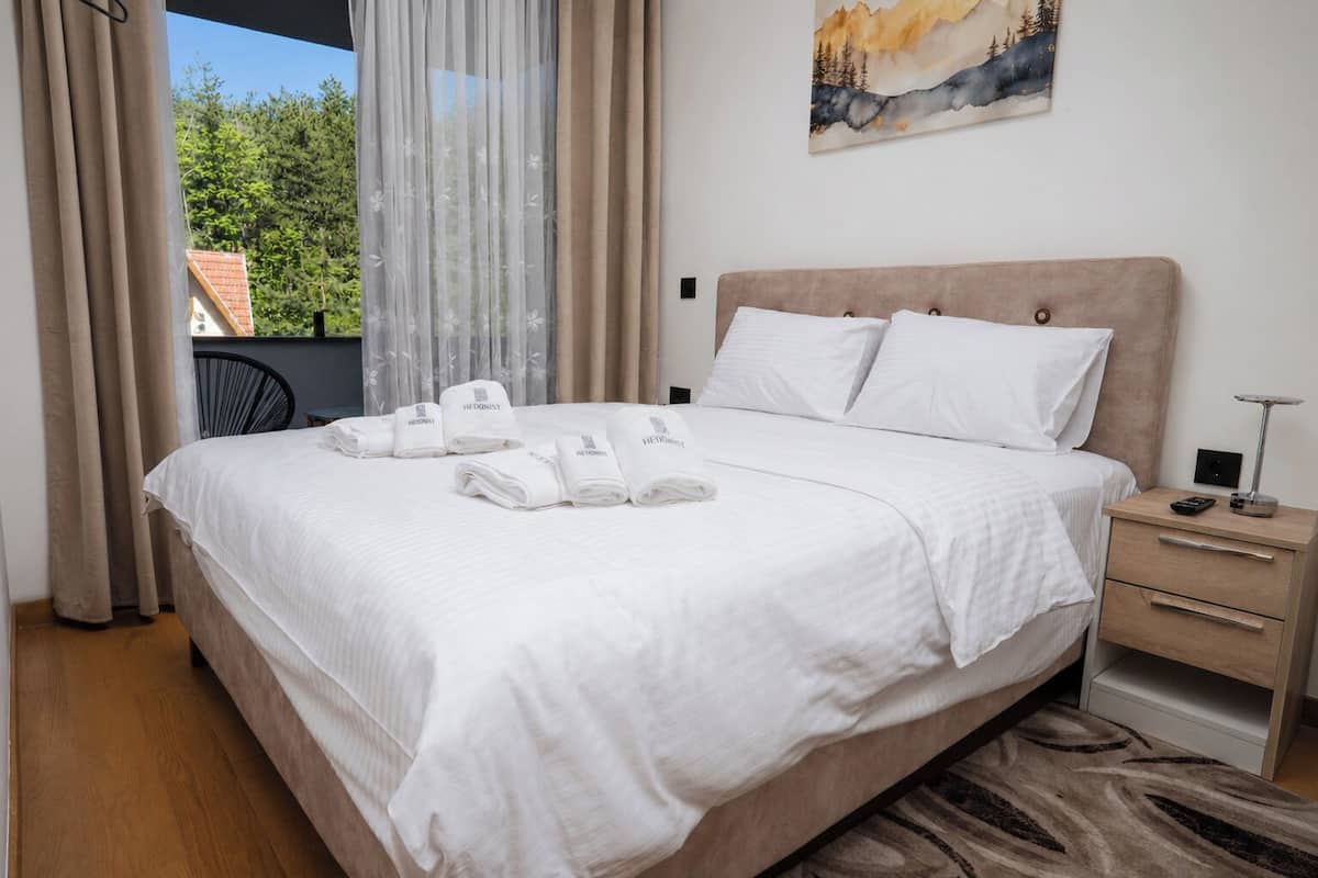 A neatly made bed with white linens and towels, a bedside table with a lamp, and a painting on the wall.