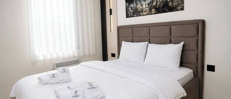 A hotel room with a large bed, white bedding, a cityscape artwork on the wall, and a window with sheer curtains.