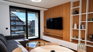 Una sala de estar moderna con un sof á, una mesa redonda, un televisor montado en una pared de madera y un ventanal de vidrio que da a un balcón exterior.