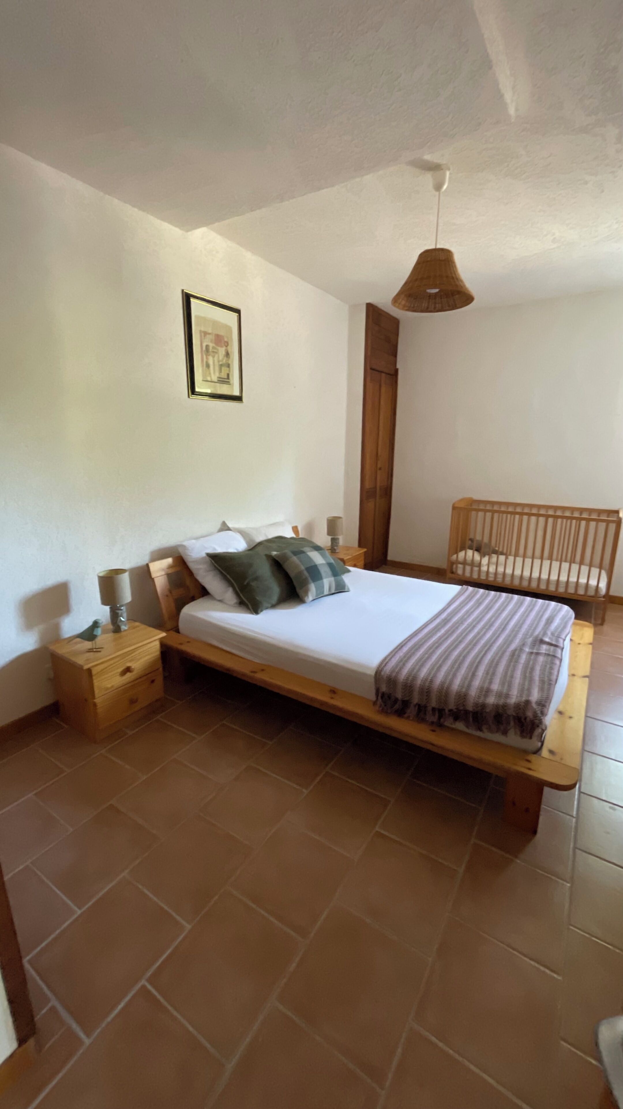 Een slaapkamer met een houten bed, nachtkastje, lamp, ledikant en hanglamp.