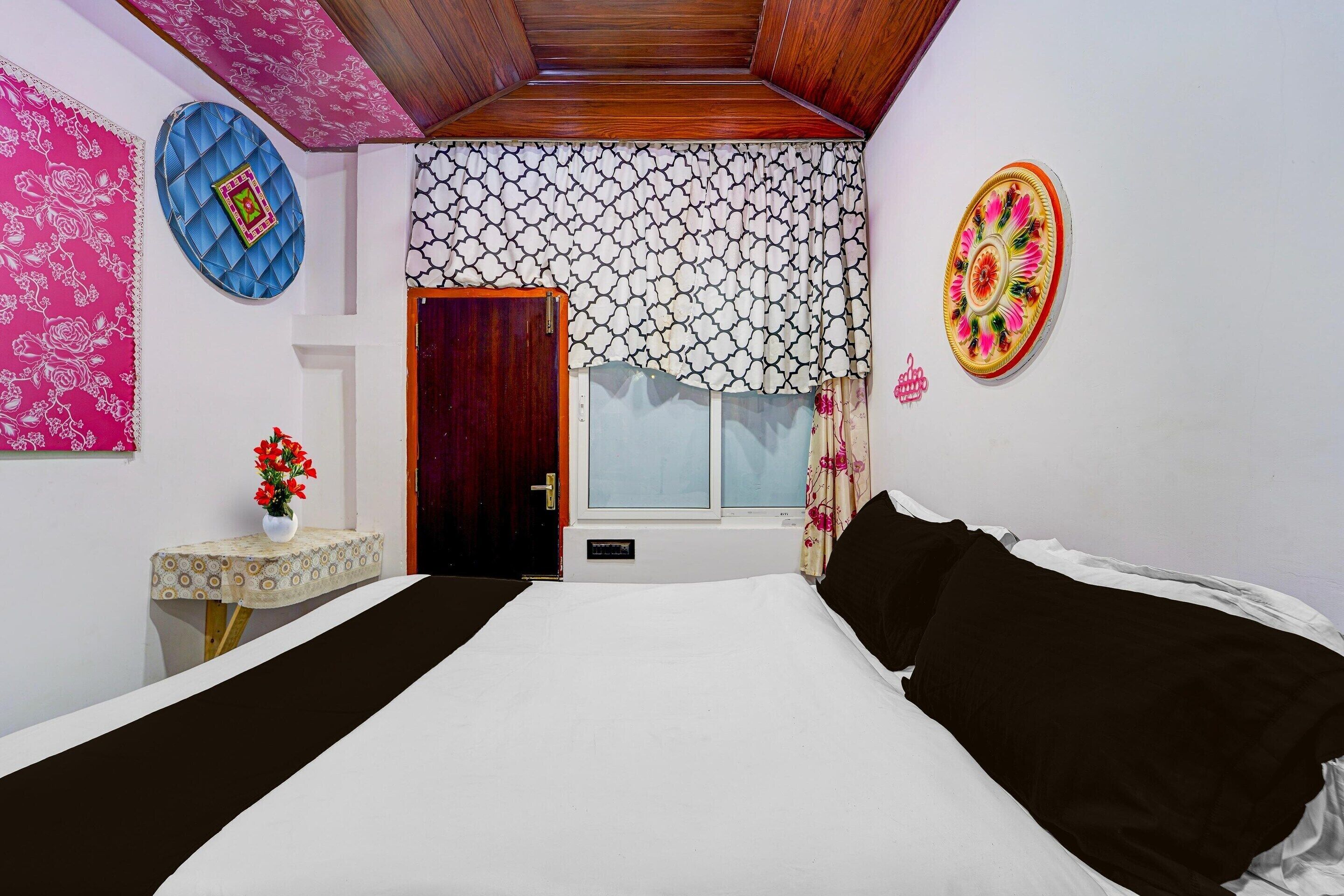 Un dormitorio con cama, una ventana con cortinas estampadas, una mesita de noche con un florero y una puerta.