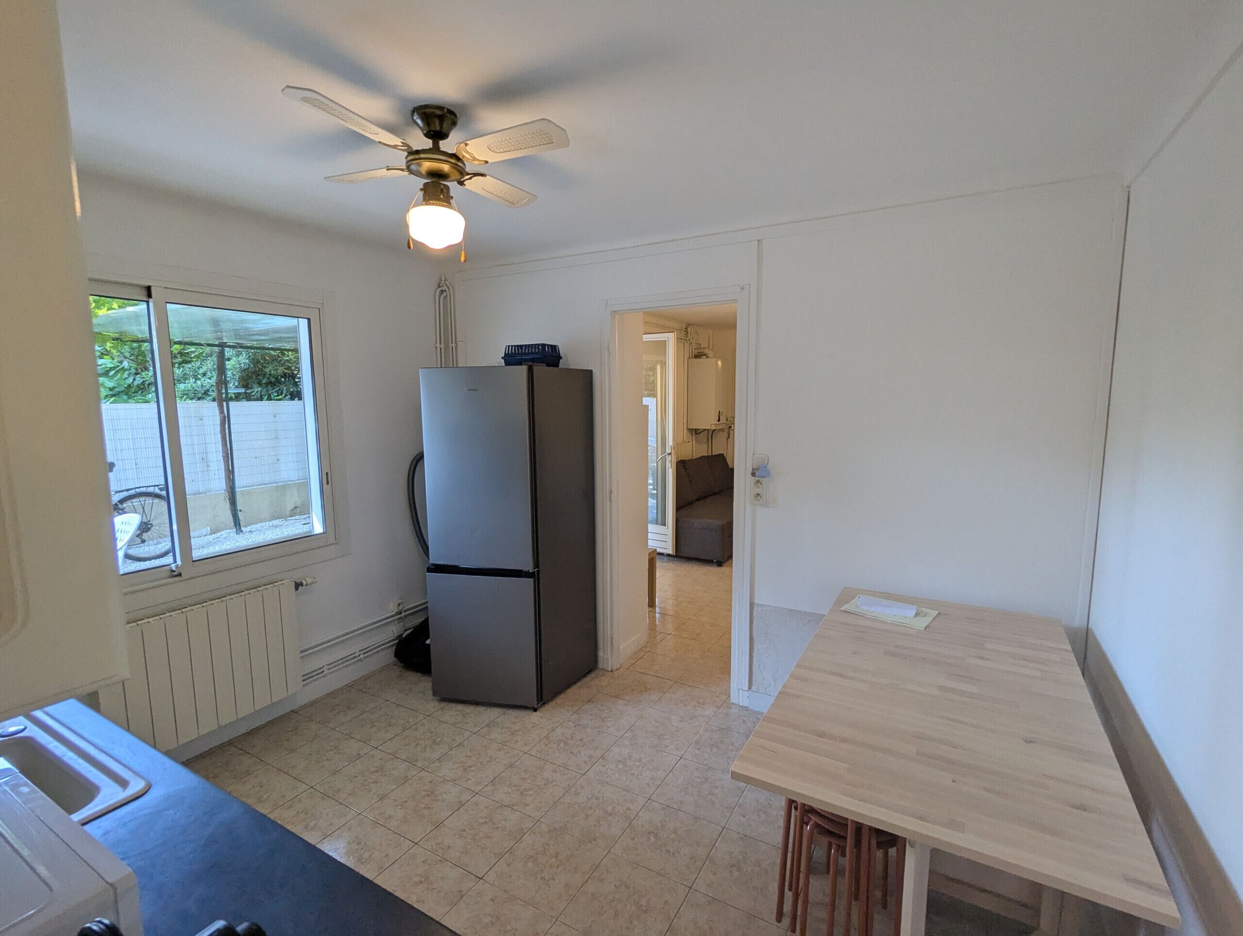 Una cocina compacta con refrigerador, ventilador de techo, ventana y puerta que da a otra habitación.