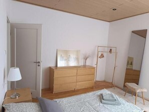 A bedroom with a wooden dresser, a bed with a patterned blanket, a bedside lamp, and a mirror. - Beautiful apartment directly at the Kurwald (Bad Lippspringe)