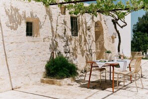 Ein rustikaler Außenbereich mit Tisch und Stühlen, einer mit Wein bewachsenen Pergola und einer weißen Wand mit Fenstern.