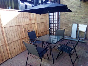 Eine Terrasse im Freien mit einem Glastisch, vier Stühlen, einem Sonnenschirm und einer Backsteinmauer.